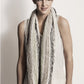 A woman with brown hair worn in a messy bun is wearing a sleeveless beige top and the Caledonia Cowl, a chunky knit infinity scarf made from Organic Cotton featuring a mix of neutral tones by Blue Sky Fibers. She stands against a plain light background and looks slightly to the side, smiling softly.