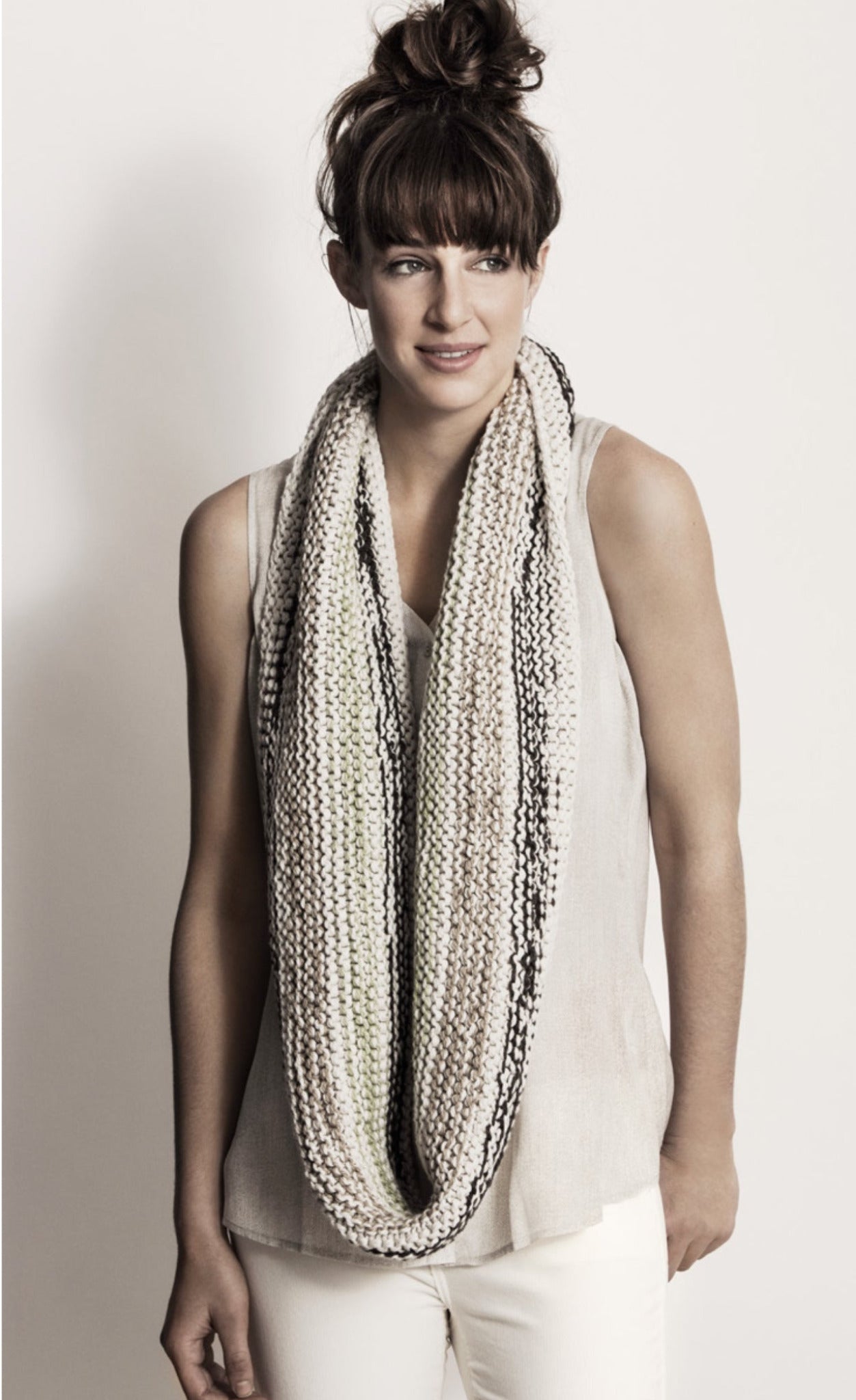 A woman with brown hair worn in a messy bun is wearing a sleeveless beige top and the Caledonia Cowl, a chunky knit infinity scarf made from Organic Cotton featuring a mix of neutral tones by Blue Sky Fibers. She stands against a plain light background and looks slightly to the side, smiling softly.