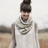 A person with long hair in a messy bun smiles while standing outdoors. They are wearing a light-colored blouse and the Caledonia Cowl from Blue Sky Fibers, a thick, knitted infinity scarf made of Organic Cotton with alternating light and dark stripes. The background is blurred, featuring a field landscape.