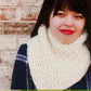 A person wearing the Eclipse Cowl by Knitting Fever / Euro Yarns— a thick, cream-colored slip stitch cowl— and a plaid jacket stands in front of a brick wall. They have long dark hair and are smiling gently at the camera.