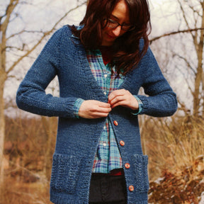 A person wearing glasses and the Tidal Cardigan by Knitting Fever / Euro Yarns, with its subtle cable details, layers it over a plaid shirt while standing outdoors. They button their cardigan among leafless trees and dry grass, suggesting an autumn or winter setting. The person looks downward while performing the task, bracing for the chilly weather.