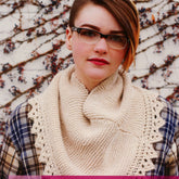 A person wearing glasses and the Boardwalk Wrap by Knitting Fever / Euro Yarns, a beige knitted cowl featuring a delicate lace edge, stands in front of a wall covered in vines with small clusters of berries. They also wear a plaid shirt. The background creates a textured and colorful contrast to their outfit.