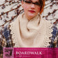 A person with short hair and glasses is wearing a plaid shirt and a beige knitted scarf with a lacy edge. The cowl and shawl, featuring garter stitch, are showcased against a vine-covered wall. Text in front of the image reads: "Boardwalk Wrap by Pam Wynne" from "The Modern Amusements Collection" by Knitting Fever / Euro Yarns.