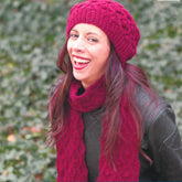 A woman with long dark hair is laughing joyfully outdoors, adorned in the chic Ramla Hat & Scarf by Knitting Fever / Euro Yarns. Her black leather jacket perfectly complements the vibrant red knit accessories against a backdrop of lush green foliage, creating a lively and harmonious scene.