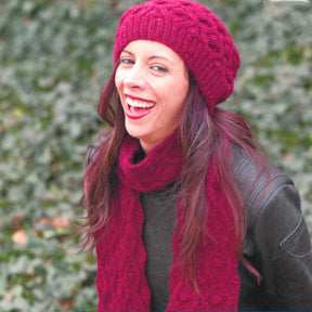 A woman with long dark hair is laughing joyfully outdoors, adorned in the chic Ramla Hat & Scarf by Knitting Fever / Euro Yarns. Her black leather jacket perfectly complements the vibrant red knit accessories against a backdrop of lush green foliage, creating a lively and harmonious scene.