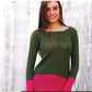 A person with long brown hair is wearing the Echo Tunic by Knitting Fever / Euro Yarns, a green, color-blocked knit sweater featuring pink accents at the bottom and sleeves. They are standing in front of a light-colored, textured wall and smiling at the camera.