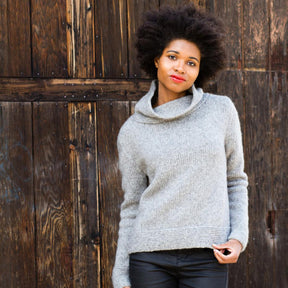 A woman with short, curly hair stands against a textured wooden door. She is wearing the Stratus Cloud Pullover sweater by Knitting Fever / Euro Yarns, made of Aran weight yarn, and black pants. She looks confidently at the camera.