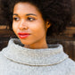 A person with curly hair stands in front of a wooden background, wearing the light gray Stratus Cloud Pullover by Knitting Fever / Euro Yarns, which features a wide cowl neck. They have a thoughtful expression and wear red lipstick.