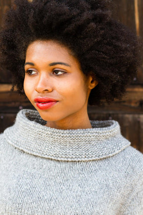 A person with curly hair stands in front of a wooden background, wearing the light gray Stratus Cloud Pullover by Knitting Fever / Euro Yarns, which features a wide cowl neck. They have a thoughtful expression and wear red lipstick.