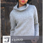 A person with medium-length curly hair is wearing a grey, knitted Stratus Cloud Pullover from Knitting Fever / Euro Yarns, made from Aran weight yarn and featuring a loose, wide collar. The individual stands in front of a rustic wooden background, with hands in the pockets of their jeans. The text on the image reads "Cloud Pullover by Pamela Wynne.