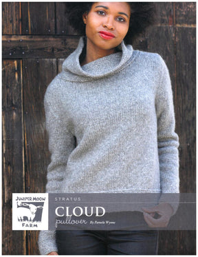 A person with medium-length curly hair is wearing a grey, knitted Stratus Cloud Pullover from Knitting Fever / Euro Yarns, made from Aran weight yarn and featuring a loose, wide collar. The individual stands in front of a rustic wooden background, with hands in the pockets of their jeans. The text on the image reads "Cloud Pullover by Pamela Wynne.