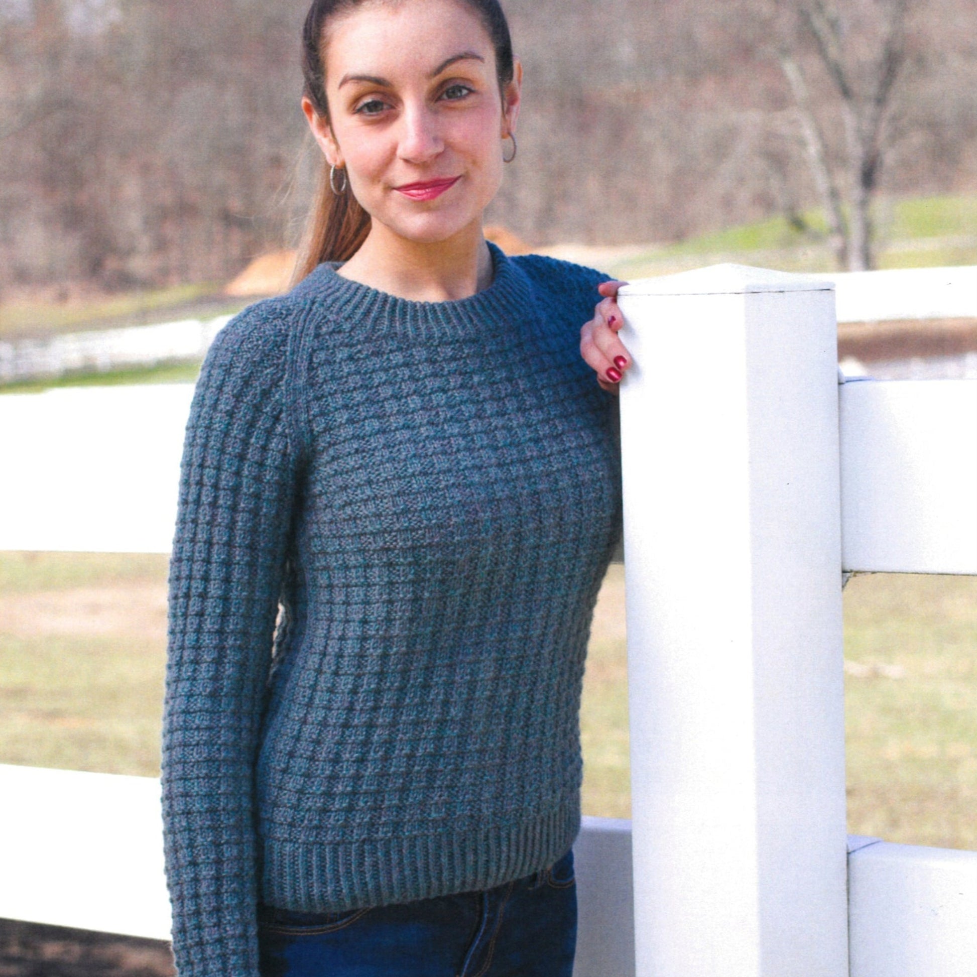 A person with long hair tied back stands outdoors on a sunny day, leaning against a white fence with one hand. They are wearing a blue Herriot Waffley Pullover from Knitting Fever / Euro Yarns, paired with blue jeans. The background features grassy fields and blurred trees. The person is smiling.