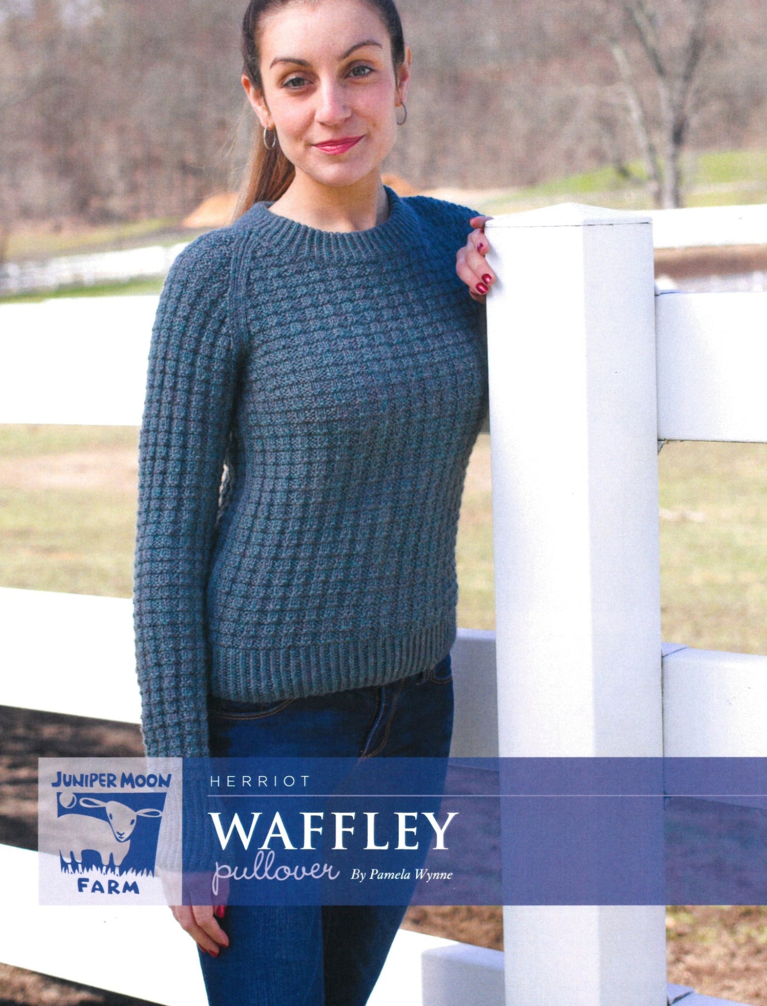 A woman with long hair stands outdoors beside a white fence, wearing a textured Herriot Waffley Pullover made from luxurious DK weight yarn in a blue-gray hue. The label at the bottom reads "Knitting Fever / Euro Yarns." The background features a grassy field and trees.