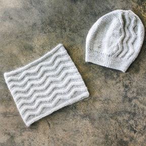 The Herriot Slalom Winter Set by Knitting Fever / Euro Yarns, featuring a light gray knitted hat and matching cowl with a wavy, textured zig-zag rib stitch pattern, is laid flat on a rustic gray concrete surface. Crafted from DK weight alpaca yarn, the hat is positioned to the right of the cowl, creating a visually appealing display of this snowy set.