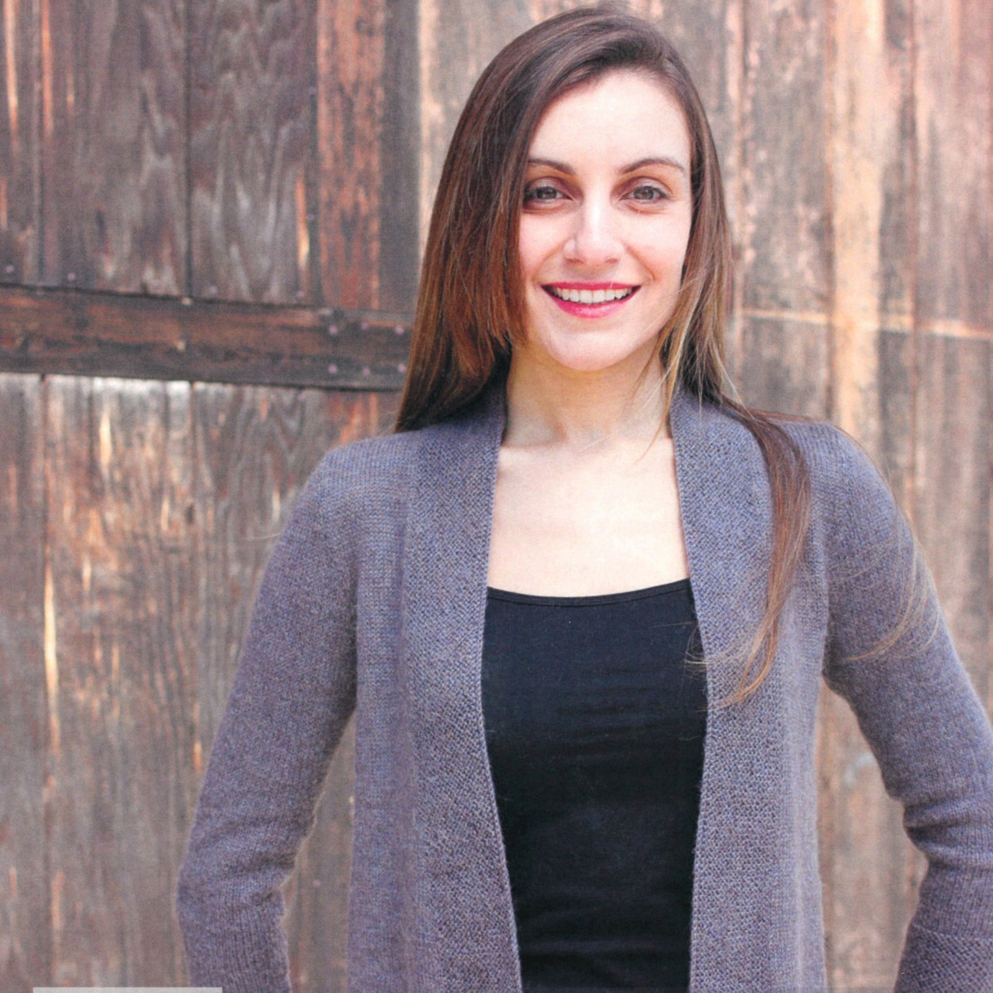 A woman with long, straight hair smiles at the camera. She is wearing a black top underneath a Herriot Fine Everyday Cardigan by Knitting Fever / Euro Yarns and standing in front of a rustic wooden backdrop.