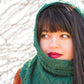 A woman with long dark hair and red lipstick wears the Herriot Fine Hedera Smoke Ring, crafted by Knitting Fever / Euro Yarns. She glances to the side against a blurred outdoor background that suggests a natural setting. Dressed in warm-colored clothing, she exudes elegance complemented by the knit cowl.