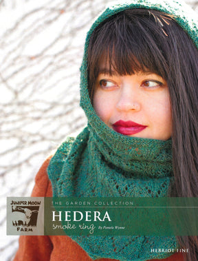 A young woman with long brown hair and deep red lipstick gazes off-camera, dressed in a green knitted cowl and cap. The text on the image reads "THE GARDEN COLLECTION Herriot Fine Hedera Smoke Ring by Pamela Wynne" and "Juniper Moon Farm HERRIOT FINE fingering weight alpaca," branded by Knitting Fever / Euro Yarns.