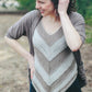 A person stands outdoors, smiling and looking to the side. They are wearing the Herriot Fine Lacebark sweater by Knitting Fever / Euro Yarns, which features a chevron pattern in neutral colors and is paired with dark pants. Their curly hair is loosely tied back, and they have one hand on their hip and the other raised to their head. The cozy Herriot Fine Lacebark sweater makes the perfect layering piece for any season.