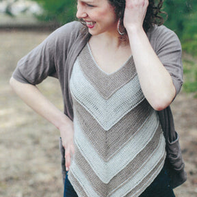 A person stands outdoors, smiling and looking to the side. They are wearing the Herriot Fine Lacebark sweater by Knitting Fever / Euro Yarns, which features a chevron pattern in neutral colors and is paired with dark pants. Their curly hair is loosely tied back, and they have one hand on their hip and the other raised to their head. The cozy Herriot Fine Lacebark sweater makes the perfect layering piece for any season.