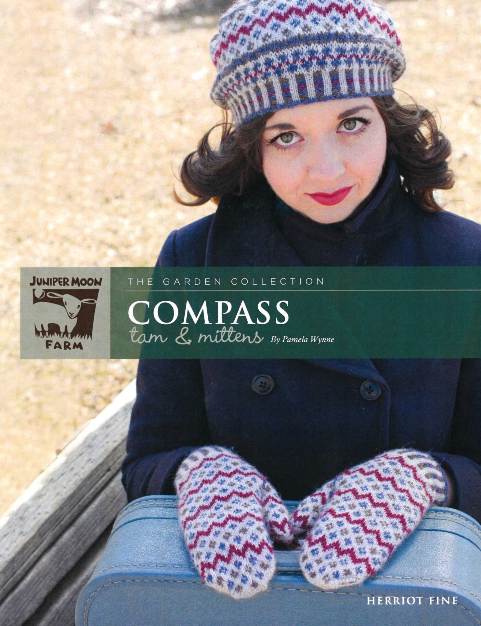 A woman wearing a knitted hat and mittens made from Knitting Fever / Euro Yarns Herriot Fine super soft alpaca, featuring a colorful zigzag pattern, holds a blue suitcase. She is dressed warmly in a dark coat, with an outdoor bench in the backdrop. The image is the cover for "Herriot Fine Compass Tam & Mittens" by Pamela Wynne.