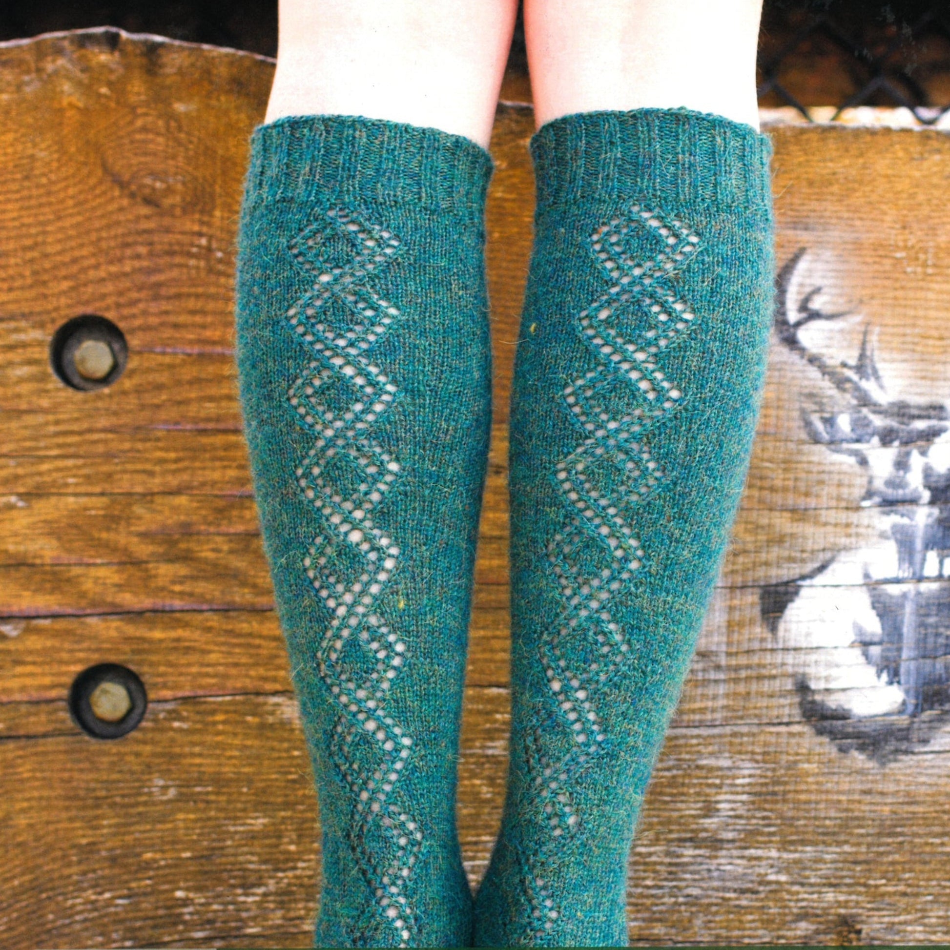 Close-up shot of a pair of legs wearing Herriot Fine Gallium Knee Socks by Knitting Fever / Euro Yarns. These teal knee-high alpaca socks feature a delicate zigzag lace pattern. The background is a wooden surface adorned with circular patterns and a partial deer head design.