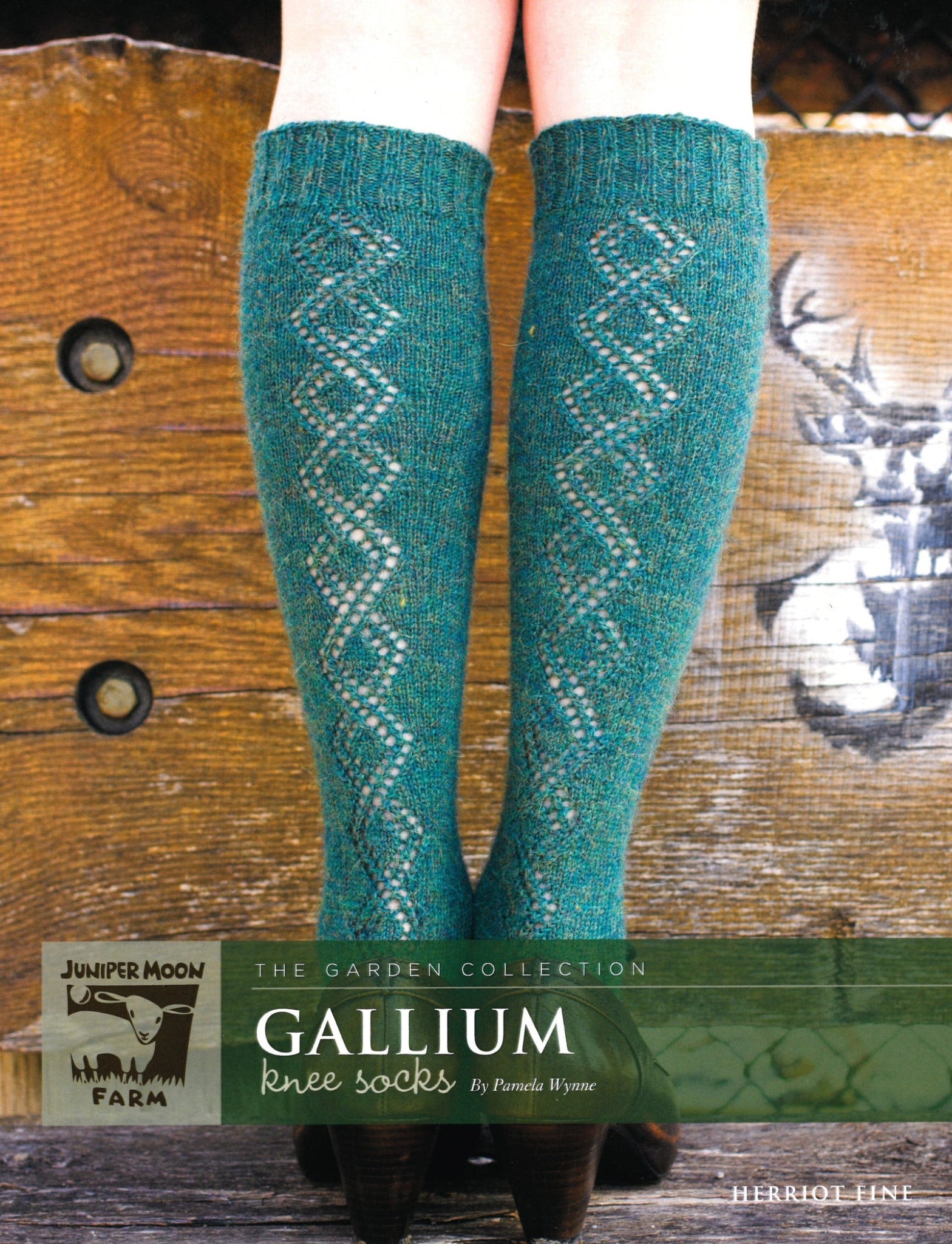 A pair of legs wearing green knee socks with a diamond pattern design. The Herriot Fine Gallium Knee Socks, from Knitting Fever / Euro Yarns, are part of The Garden Collection by Juniper Moon Farm and designed by Pamela Wynne. The background features a rustic wooden wall with an artistic mural.