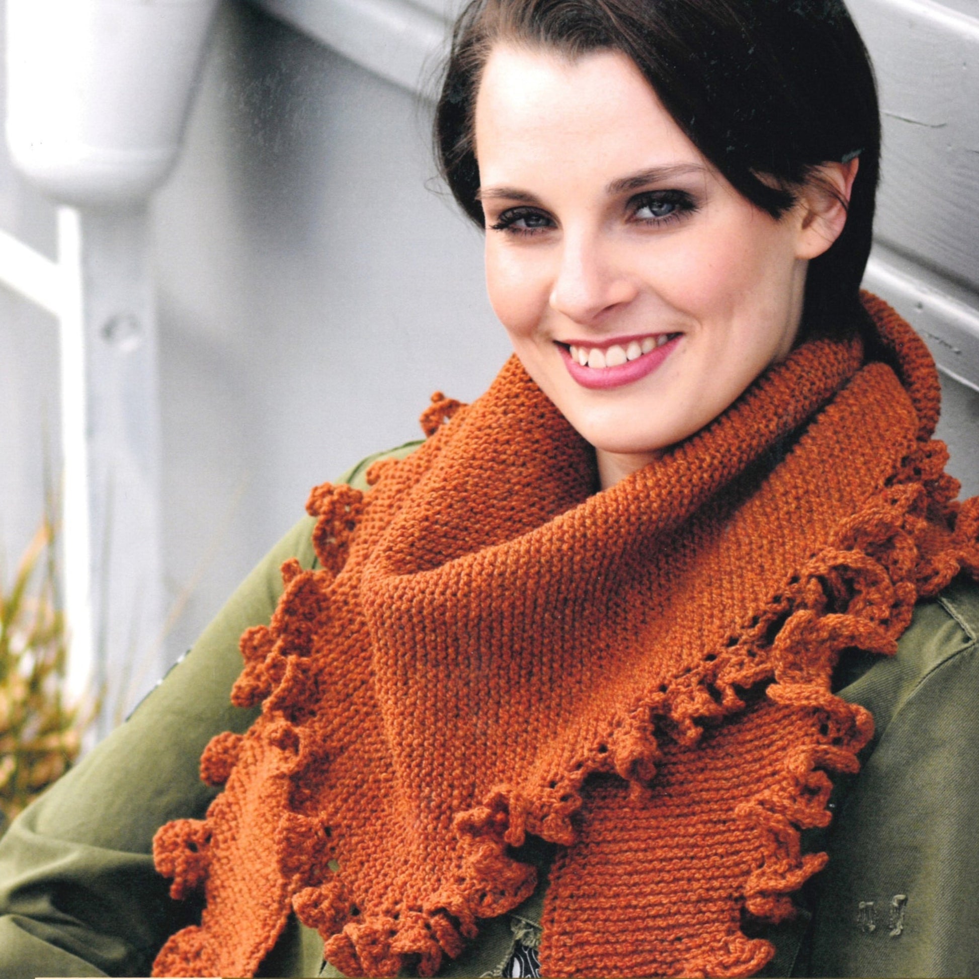 A person with short dark hair smiles while wearing the Matilda Shawl, a knitted piece from Knitting Fever / Euro Yarns, in a rust color made from DK weight yarn with ruffled edges. The person is dressed in an olive green jacket, and behind them is a gray wall with some vegetation partially visible.