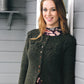 A woman smiles while standing against a white wall. She is wearing a Knitting Fever / Euro Yarns Thjorven Jacket in dark green, adorned with a row of brown buttons down the front, and a floral dress underneath. Her long, light brown hair is down, and she appears to be outside on a porch.