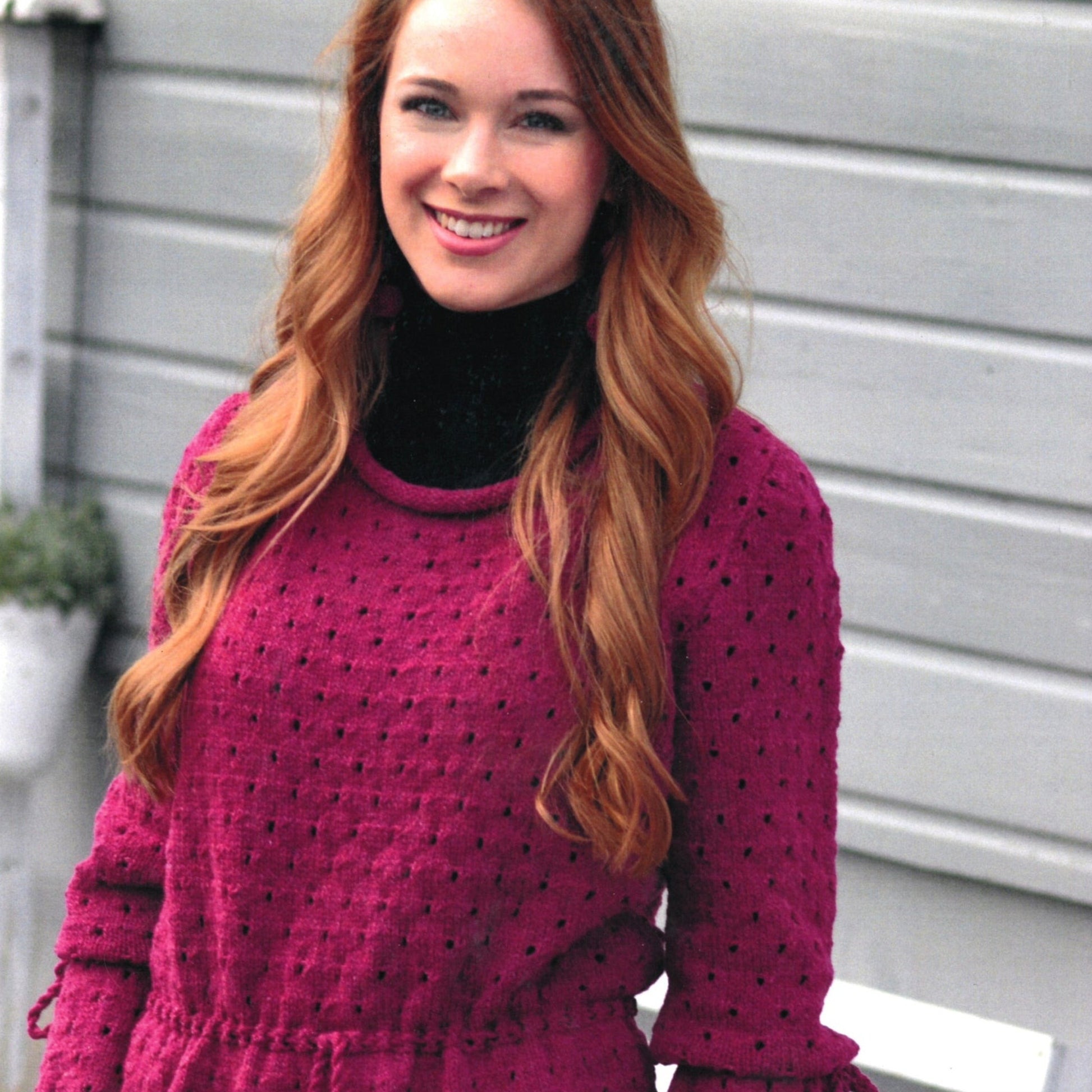 A woman with long, wavy auburn hair smiles while posing outdoors. She is wearing a magenta Svenja Sweater from Knitting Fever / Euro Yarns over a black turtleneck. The background features a gray wooden wall and a potted plant. The eyelet pattern on her sweater adds an elegant touch to her outfit.