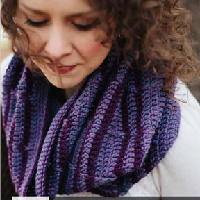 A person with curly hair and closed eyes is shown wearing a chunky, stripey Herriot Trellised Wisteria Crochet Cowl by Knitting Fever / Euro Yarns. The cozy piece appears perfect for a winter day, and the person is dressed in a light-colored top, enhancing the warm feel. The background is blurry, drawing focus to the individual and their stylish scarf.