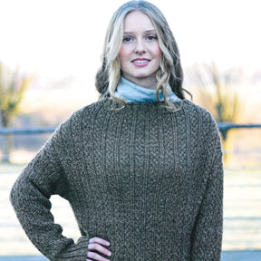 A woman with long, blonde hair smiles at the camera. She is wearing a knitted Herriot Metta Sweater by Knitting Fever / Euro Yarns, perfect for a weekend adventure, and a blue-gray scarf. The background is a blurred outdoor scene with trees and a fence.