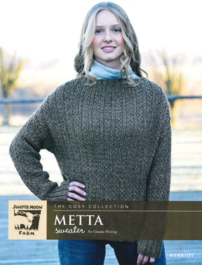 A woman stands outdoors wearing the versatile Herriot Metta Sweater, a brown knitted pullover with a ribbed pattern. The background includes a wooden fence and trees adorned with autumn foliage. The text on the image reads, "THE COSY COLLECTION," "Knitting Fever / Euro Yarns," "METTA sweater by Claudia Wersing," and "HERRIOT.