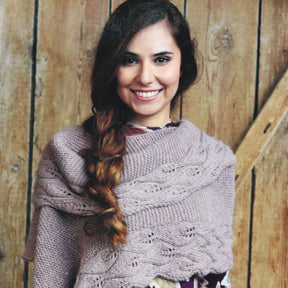 A woman with long, braided hair smiles warmly at the camera. She is wrapped in a cosy Herriot Odveig Wrap from Knitting Fever / Euro Yarns, featuring intricate patterns over a patterned top. The background features wooden planks, adding a rustic touch to the scene.