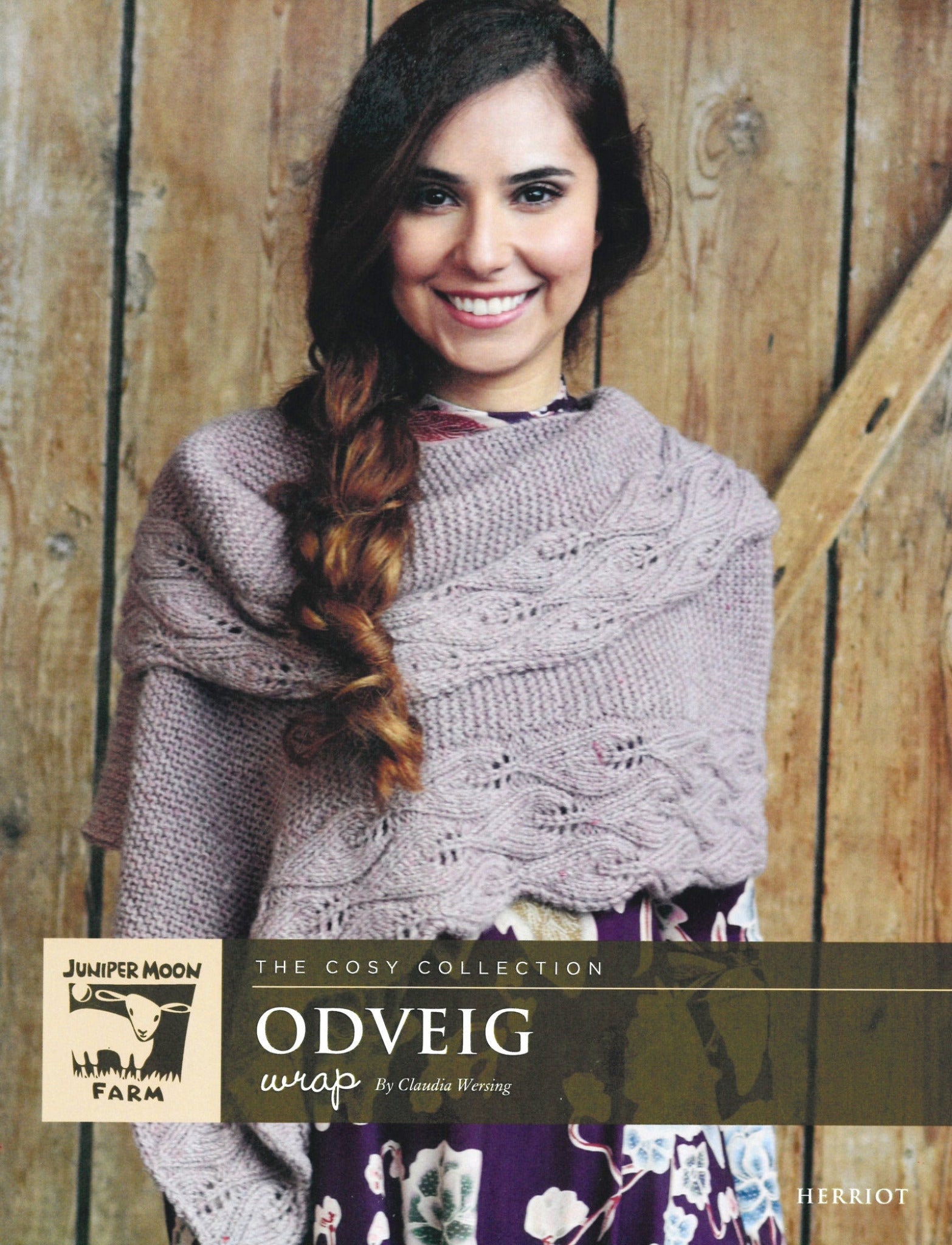 A woman with long brown hair in a braid smiles at the camera, wearing a Herriot Odveig Wrap over a floral dress. She stands against a wooden backdrop. The text on the image reads: "Juniper Moon Farm," "The Cosy Collection," and "Herriot Odveig Wrap by Claudia Wersing" from Knitting Fever / Euro Yarns." The knit wrap looks exceptionally cosy.