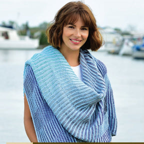 A smiling woman with short brown hair stands outdoors near a body of water. She is wearing a white top and the Cumulus Rainbow Zeynep Wrap by Knitting Fever / Euro Yarns, which features blue and turquoise striped Cumulus yarn draped over her shoulders. The background includes blurred boats and water.