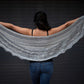 A person with long dark hair is standing with their back facing the camera, arms outstretched to show off a large Cumulus Sasha Crescent Shawl from Knitting Fever / Euro Yarns, featuring a lace border and intricate patterns. They are wearing a black top and blue jeans, against a dark background.