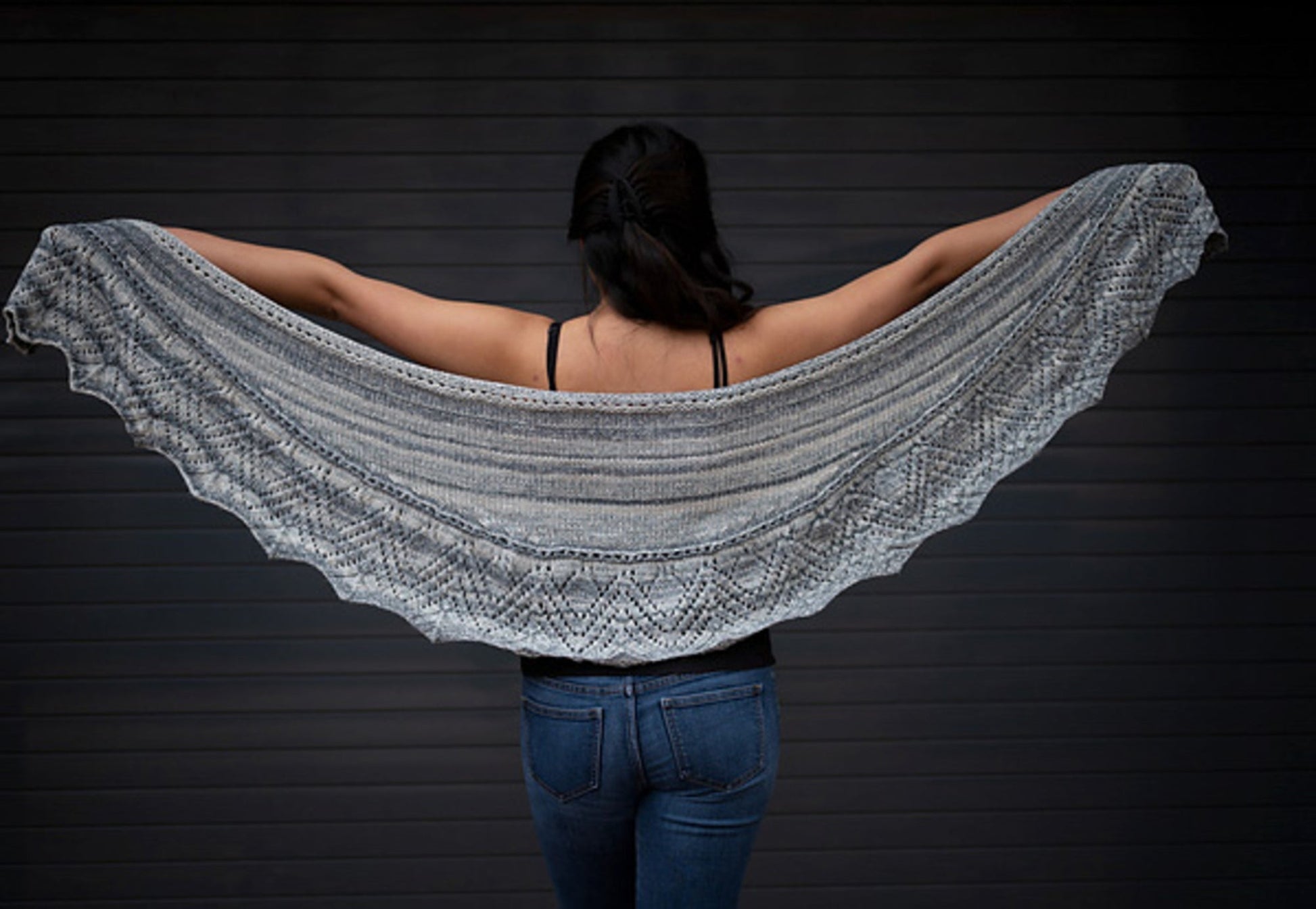 A person with long dark hair is standing with their back facing the camera, arms outstretched to show off a large Cumulus Sasha Crescent Shawl from Knitting Fever / Euro Yarns, featuring a lace border and intricate patterns. They are wearing a black top and blue jeans, against a dark background.