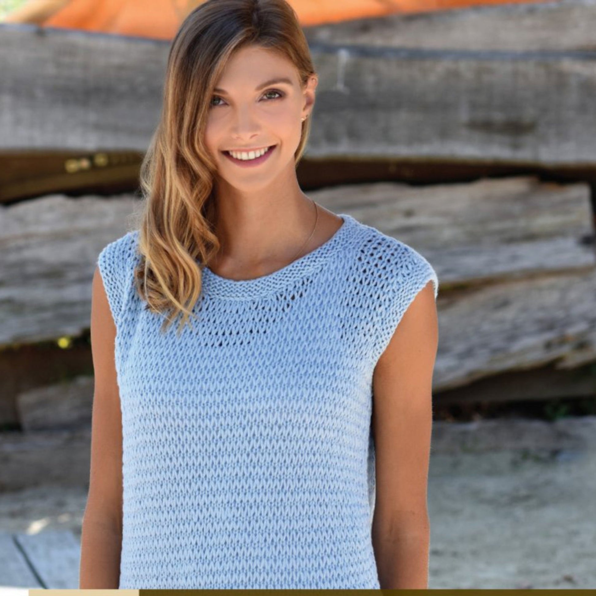 A woman with long blonde hair smiles while wearing the Cumulus Dorothea Tank Top, a light blue, sleeveless knit creation by Knitting Fever / Euro Yarns, perfect as a swimsuit cover-up. She stands outdoors with a rustic wooden fence in the background.