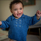 A joyful baby with curly hair is wearing a cozy Cumulus Marlowe Baby Jacket from Knitting Fever / Euro Yarns, standing up and smiling exuberantly with arms outstretched. The background is softly blurred, drawing focus to the baby's happy expression.