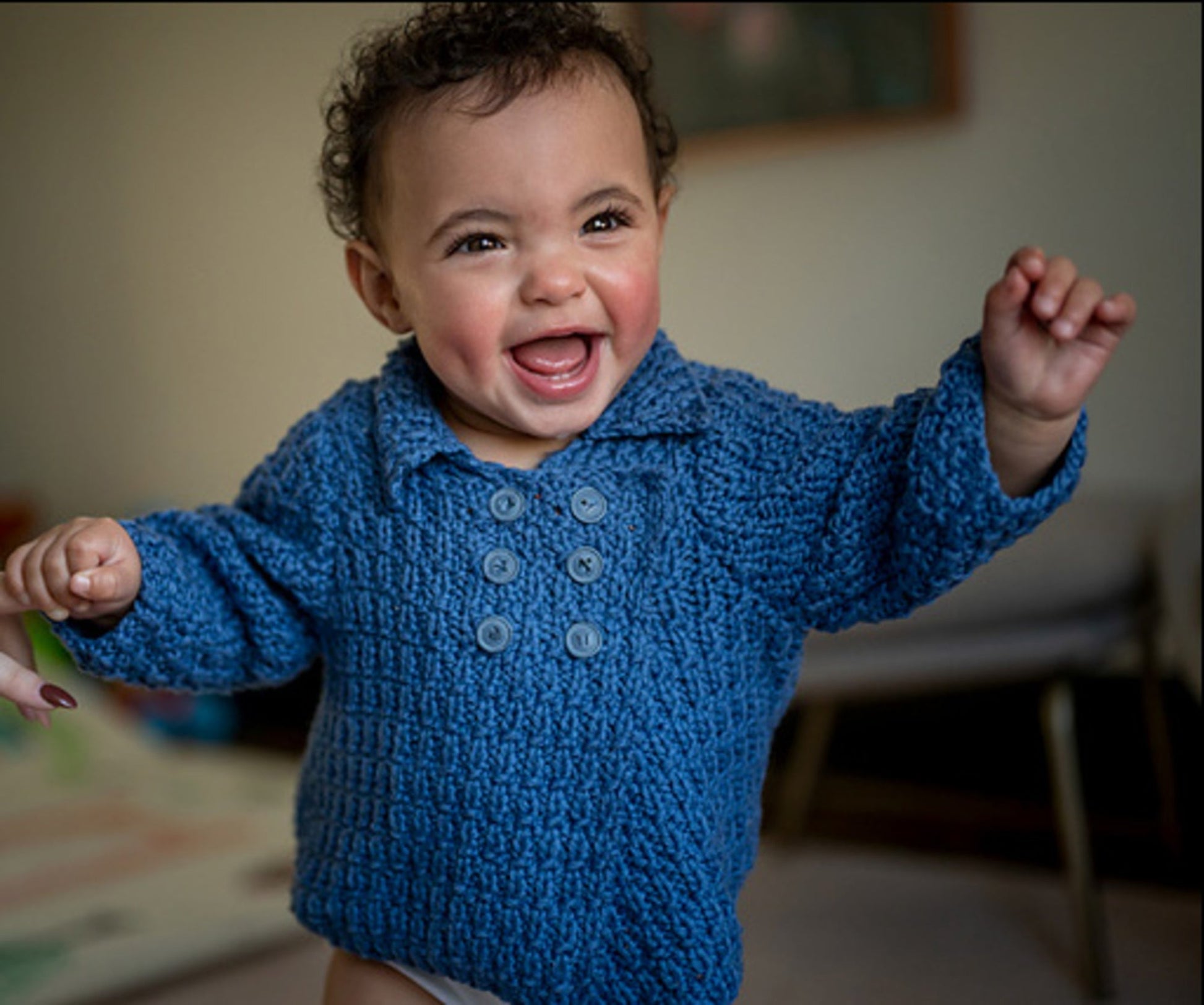 A joyful baby with curly hair is wearing a cozy Cumulus Marlowe Baby Jacket from Knitting Fever / Euro Yarns, standing up and smiling exuberantly with arms outstretched. The background is softly blurred, drawing focus to the baby's happy expression.