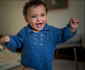 A joyful baby with curly hair is wearing a cozy Cumulus Marlowe Baby Jacket from Knitting Fever / Euro Yarns, standing up and smiling exuberantly with arms outstretched. The background is softly blurred, drawing focus to the baby's happy expression.