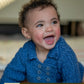 A happy toddler with curly hair smiles widely while looking away from the camera. The child is wearing a cozy Cumulus Marlowe Baby Jacket in blue, designed by Knitting Fever / Euro Yarns, featuring buttons down the front. The background is slightly blurred, drawing focus to the child's joyful expression.