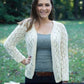 A woman is standing outdoors on grass with trees in the background. She is smiling and wearing a grey tank top, a light cotton Cumulus Faros Cardigan by Knitting Fever / Euro Yarns with a lace pattern, and dark jeans with a brown belt. One hand is on her hip, and she is looking into the camera.