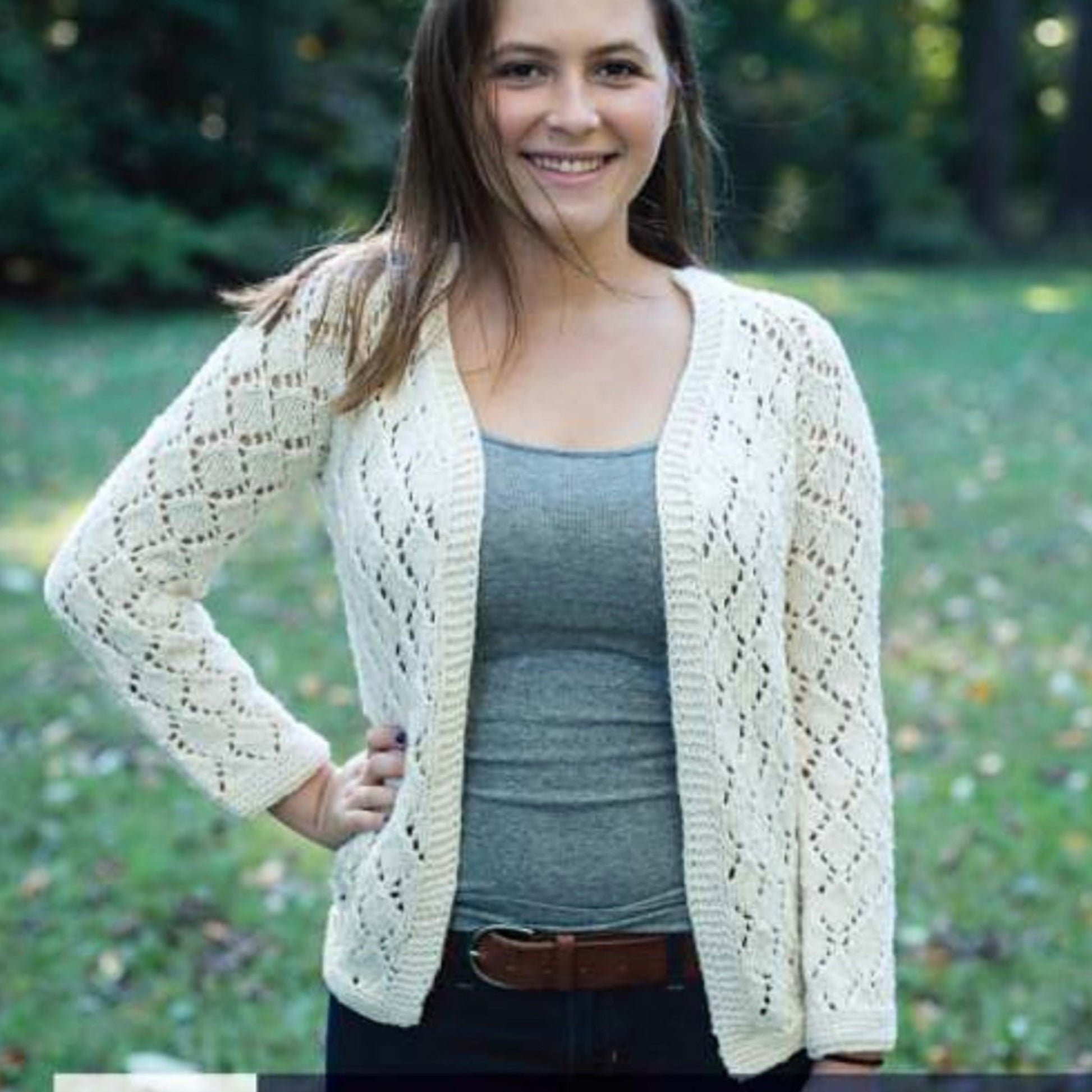 A woman is standing outdoors on grass with trees in the background. She is smiling and wearing a grey tank top, a light cotton Cumulus Faros Cardigan by Knitting Fever / Euro Yarns with a lace pattern, and dark jeans with a brown belt. One hand is on her hip, and she is looking into the camera.