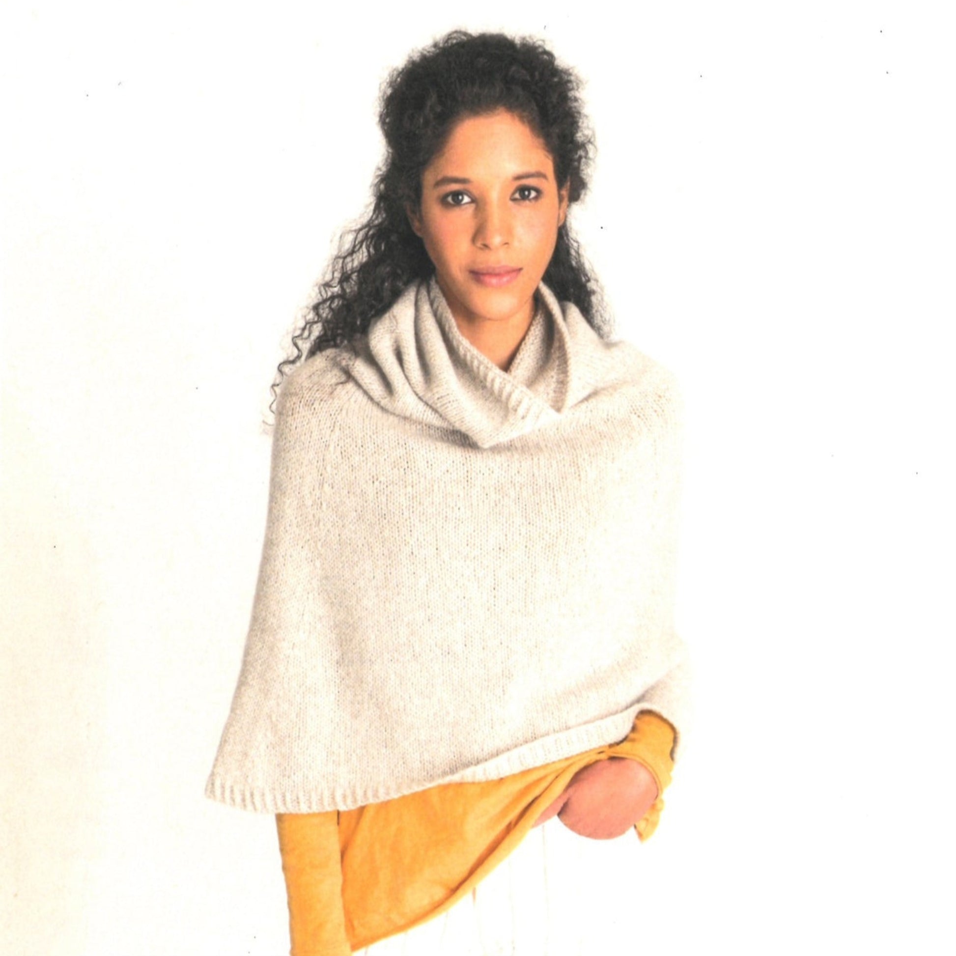 A woman with curly hair wears a Blue Sky Fibers Princeton Capelet in cream over a mustard yellow sweater. She has her left hand casually tucked into her pocket and stands against a plain white background, looking calmly at the camera. This versatile piece is perfect for any occasion.