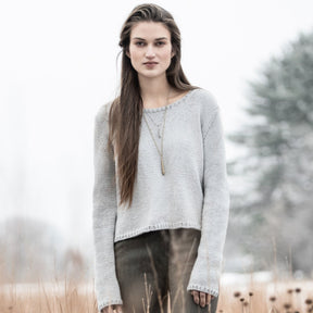 A woman with long brown hair stands in a field of tall grass, wearing a Blue Sky Fibers Spring Hill Sweater and a necklace. The background is soft and hazy, with trees and a cloudy sky. The overall tone is muted and serene.