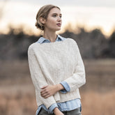 A woman stands outside, gazing into the distance. She has blonde hair tied back and is wearing the Shady Hollow Sweater by Blue Sky Fibers, a cream-colored, textured sweater with a wide neckline over a light blue shirt. The background is an out-of-focus natural setting with trees and grass under a cloudy sky.