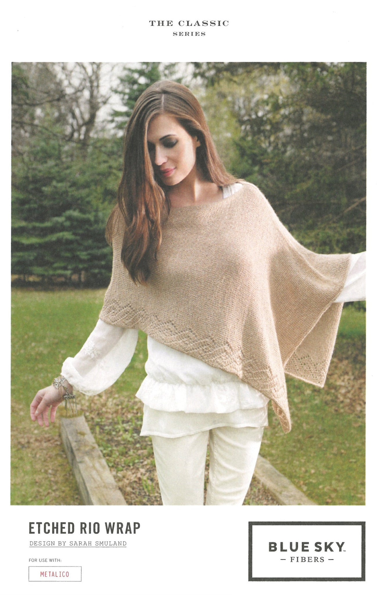 A woman stands outdoors wearing a beige knitted wrap with a delicate lace panel over a white top and pants. The wrap, known as the Etched Rio Wrap, is part of "The Classic Series" by Blue Sky Fibers and is designed by Sarah Smuland. Trees and greenery provide a natural background.