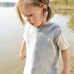 A young child with blonde hair in pigtails is standing near a calm body of water. The child is wearing the Cameron Hoodie by Blue Sky Fibers, a cozy gray knitted sweater over an organic cotton vest, and is looking downwards with a thoughtful expression. The background shows reeds and the lake in soft focus.