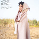 A happy child stands outdoors, wrapped in an extra soft, hooded "Georgie Blanket" by Blue Sky Fibers, smiling and looking to the side. The beige handmade heirloom, made with organic cotton and featuring green edges, exudes comfort. In the background, grassy fields stretch under a hazy sky. Text reads "GEORGIE BLANKET ORGANIC COTTON COLLECTION.